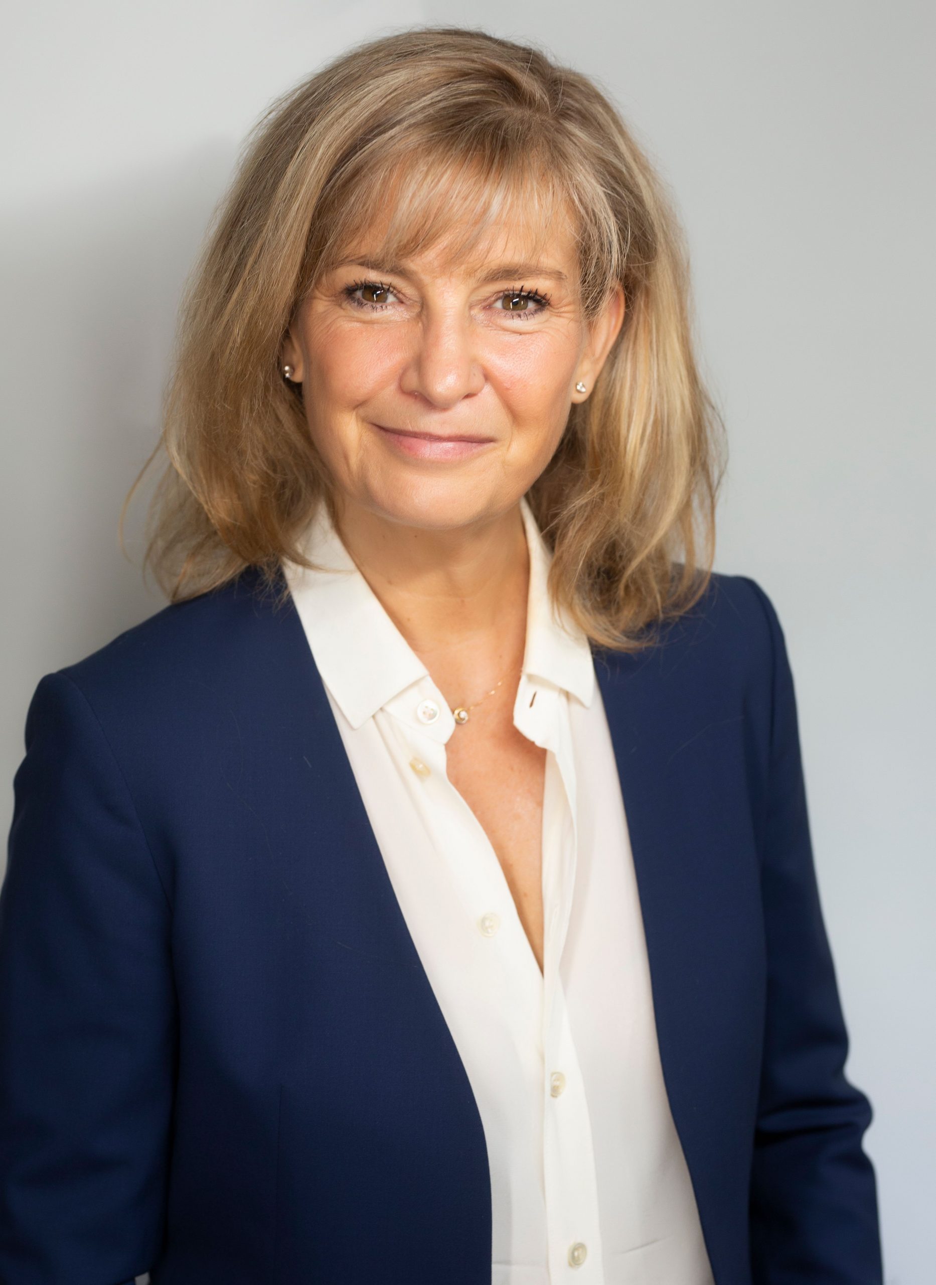 Jessica Mosher standing against a beige wall. Woman with dark blonde hair, smiling and wearing a white shirt and blue blazer.