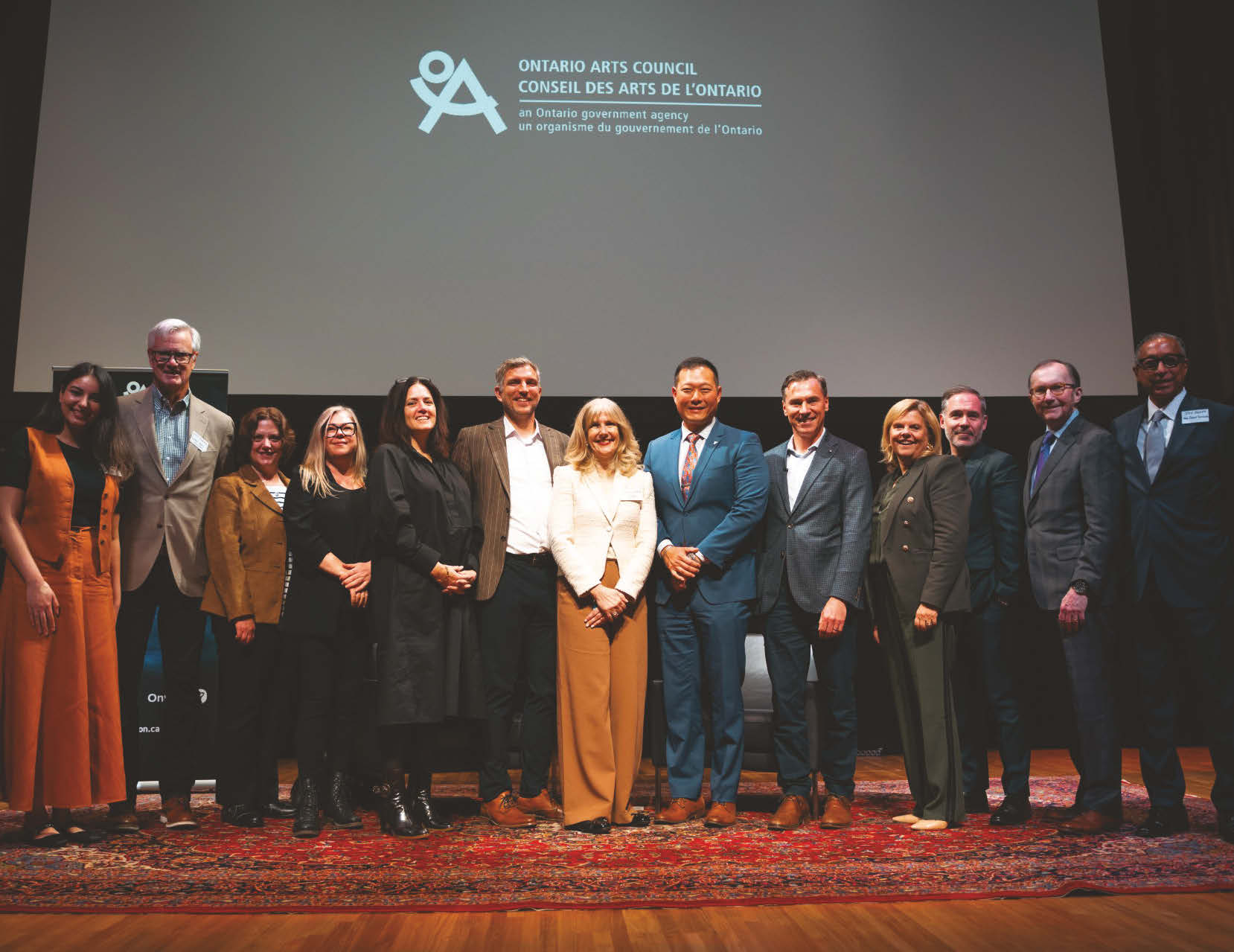 Photo of 13 people standing side by side under an Ontario Arts Council logo